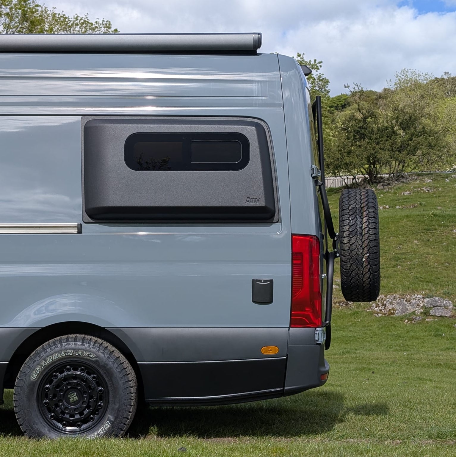 Side and rear view of a light gray camper van parked outdoors on grass under a blue sky with clouds, showing ADV side pod.